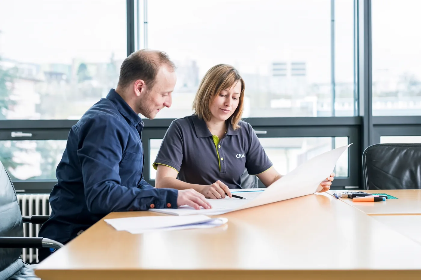 Two colleagues reviewing technical documents and a laptop screen in a bright meeting room